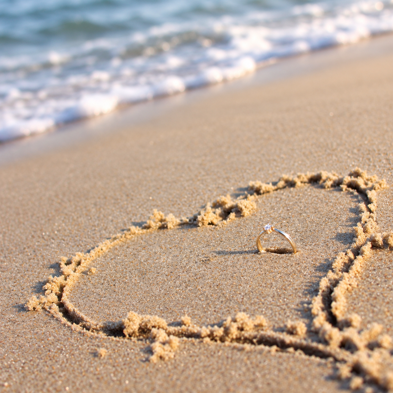 Engagement ring placed right in center of a heart drawn on a beach sand.