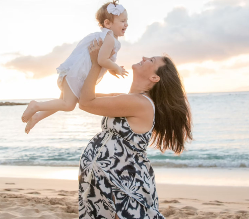 A pregnant woman lifts her young daughter into the air during Maui photoshoot for families. The sun sets in the background on the water.