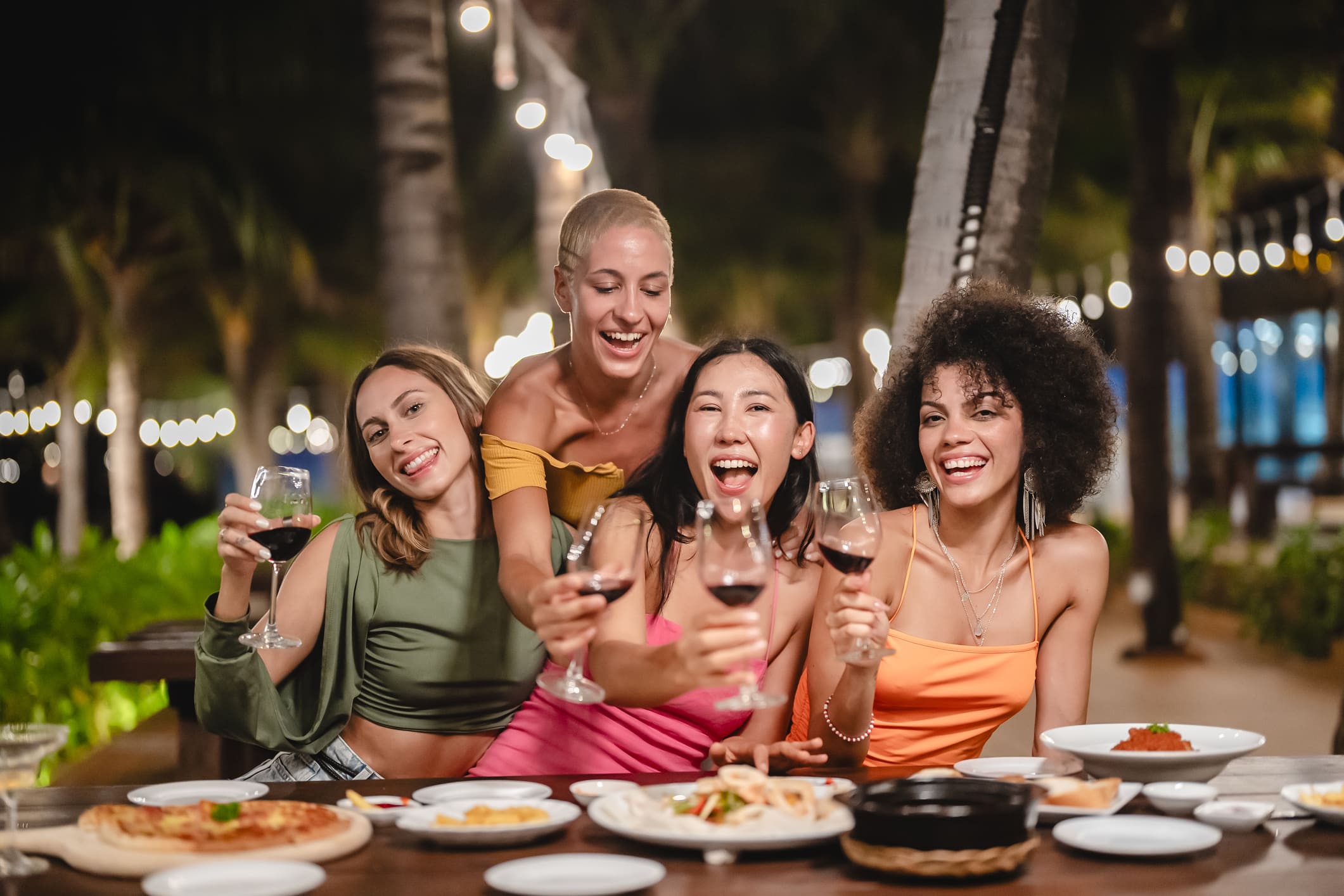Group of happy friends sharing laughs and drinks at an outdoor dinner party in Maui, under palm trees and string lights.